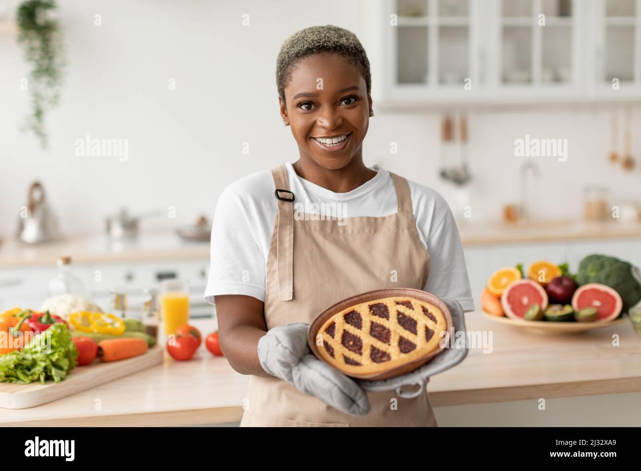 Smiling pretty millennial african american woman in apron showing tasty pie in minimalist kitchen interior Stock Photo