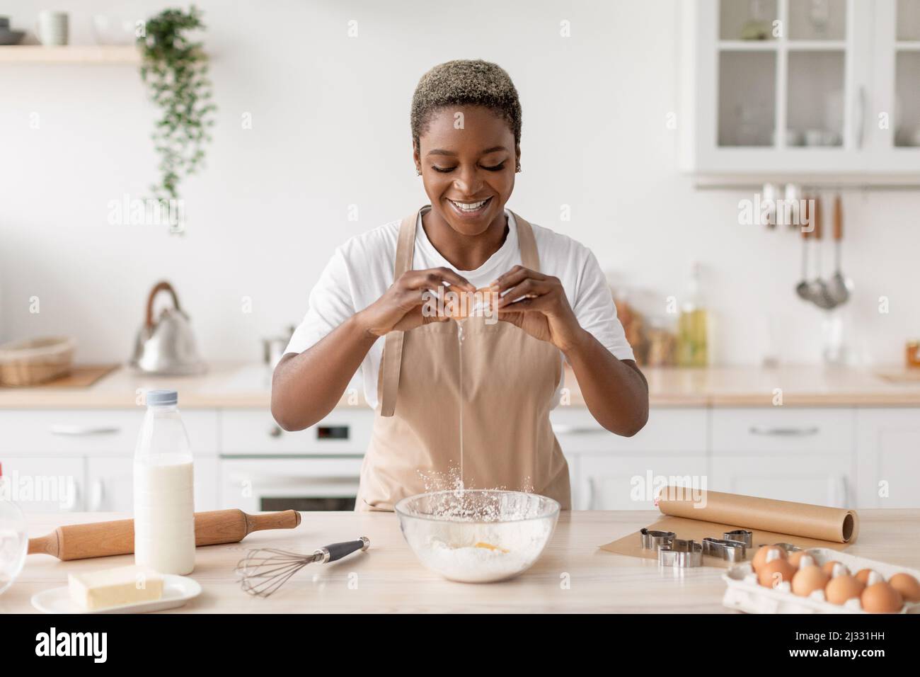 Cheerful millennial african american lady in apron breaks egg preparing dough in minimalist kitchen interior Stock Photo