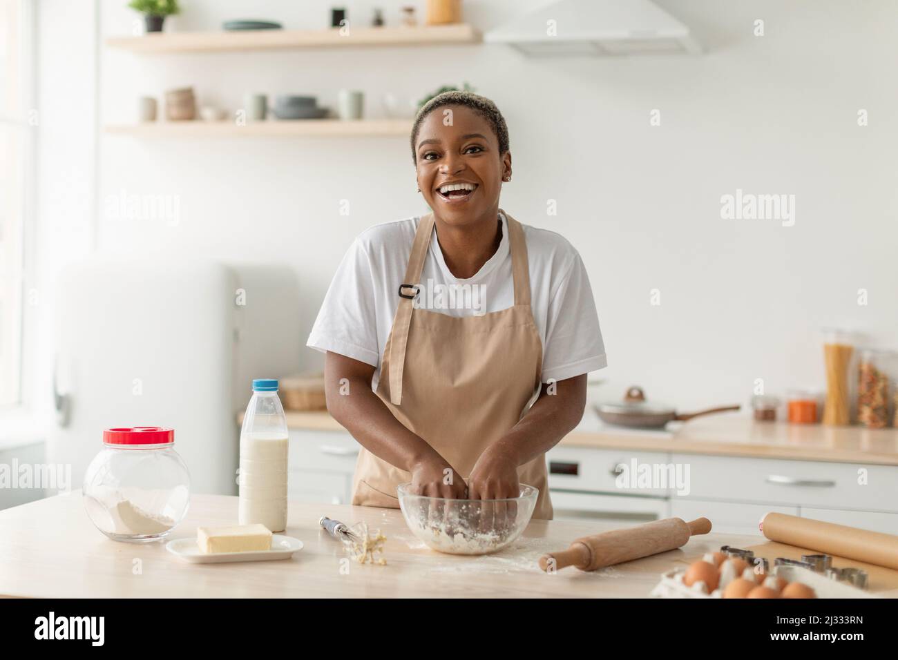 Laughing young african american lady in apron making pie dough in minimalist kitchen interior Stock Photo