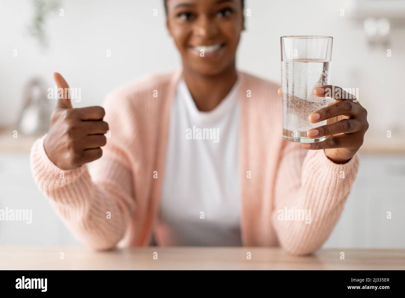 Happy young african american lady hold glass of clean water and show thumb up in minimalist kitchen Stock Photo
