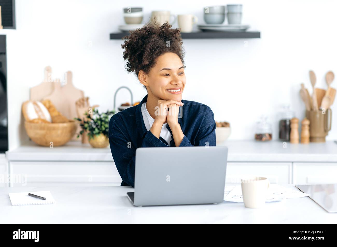 Portrait happy pretty African American girl, stylishly dressed, with curly hair, freelancer, student, designer, work from home uses laptop, sit in the kitchen, looking away, thinking, dreaming, smile Stock Photo