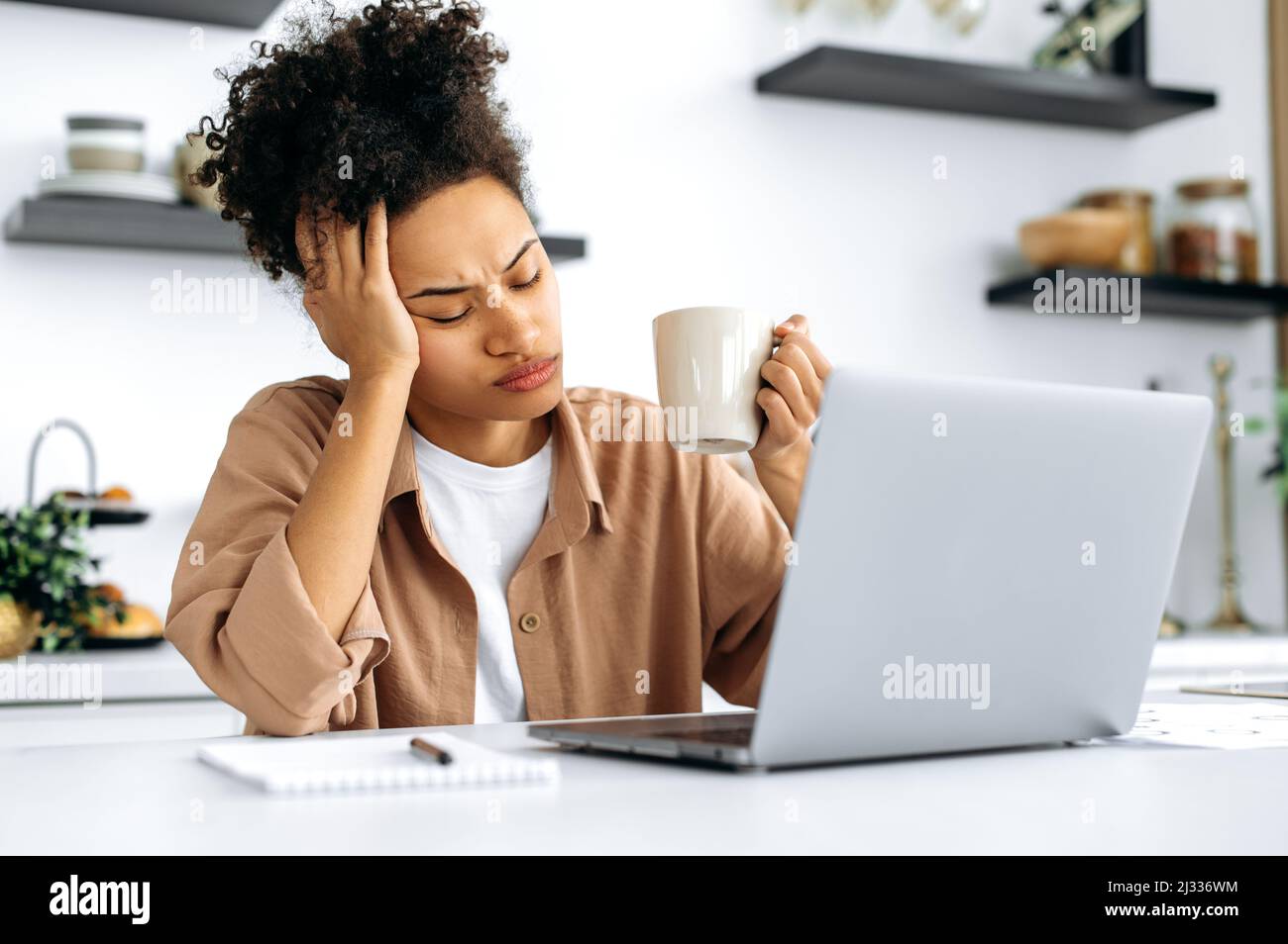 Bored african american girl, freelancer, designer, working remotely from home, tired of boring online work, suffering from chronic fatigue or overwork, holds a cup of coffee in her hand, falls asleep Stock Photo