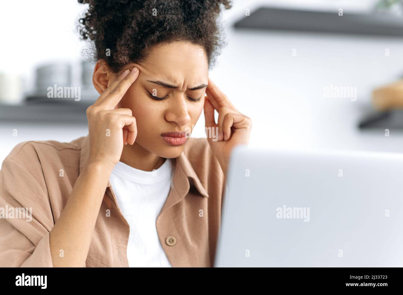 Close-up of a tired african american exhausted girl, experiencing fatigue from work, overwork, suffering from headache, migraine, massages her temples, closed her eyes, sits at a laptop, needs rest Stock Photo