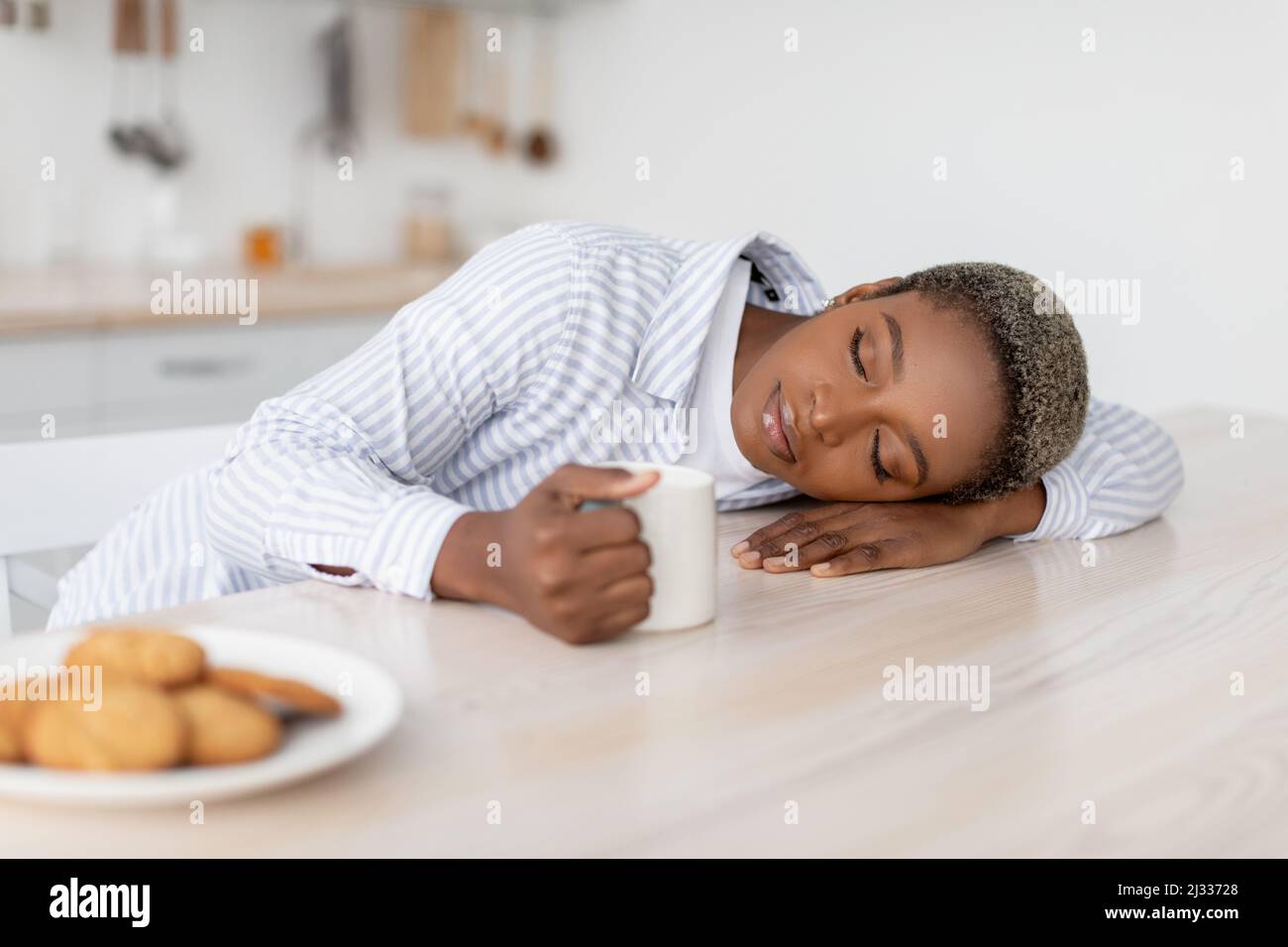 Tired young african american female with coffee cup sleeping on table in modern minimalist white kitchen interior Stock Photo
