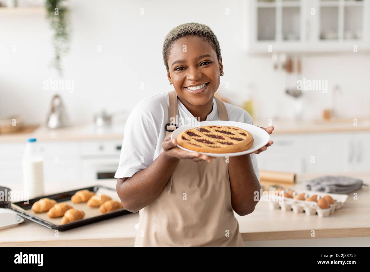 Cheerful happy funny millennial african american female cook in apron showing fresh pie in white kitchen interior Stock Photo