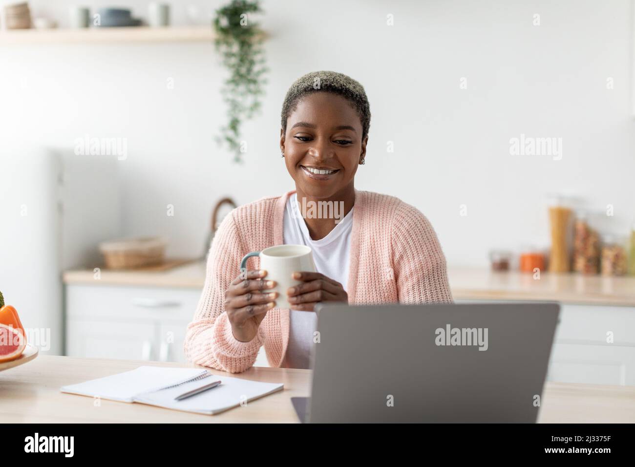 Happy millennial african american woman hold cup and work with laptop in modern minimalist kitchen interior Stock Photo