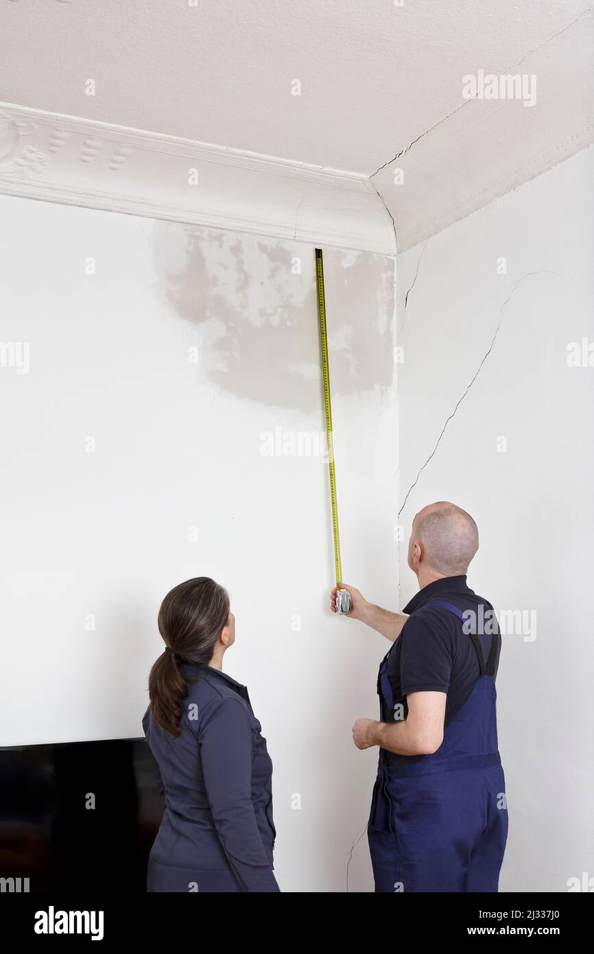 Janitor or building caretaker measuring the extent of a big water stain on the cracked wall of an old building, female tenant watching. Stock Photo