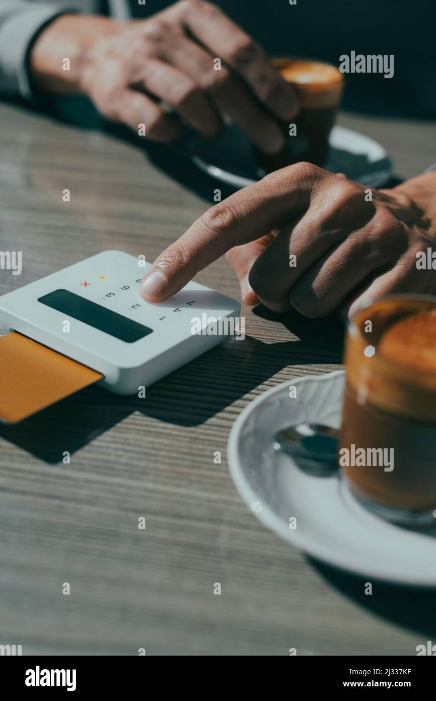 a young man, sitting at a table of a sidewalk cafe, inserts the PIN of his credit card on the keypad of a wireless payment terminal, to pay the bill Stock Photo