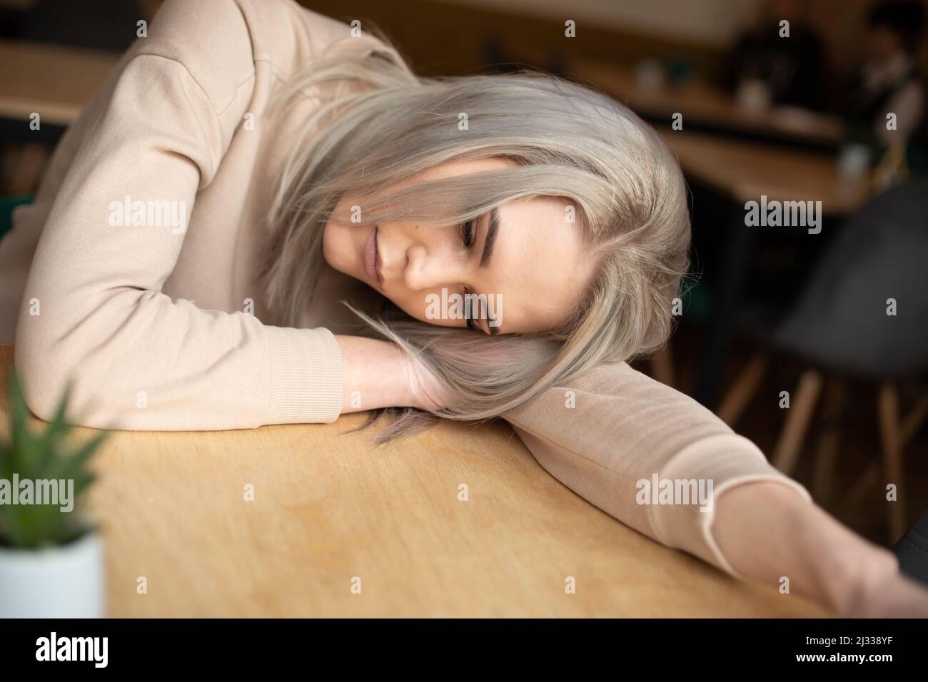 Portrait of young attractive pensive woman with long grey hair with make-up in sweatshirt laying head on hand on table. Stock Photo
