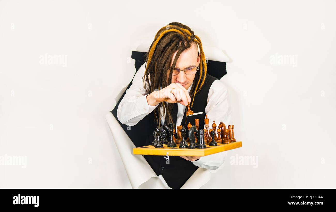 Young man in glasses with dreadlocks plays chess in hole of white background. Handsome male in white shirt, black vest and tie playing in logical boar Stock Photo