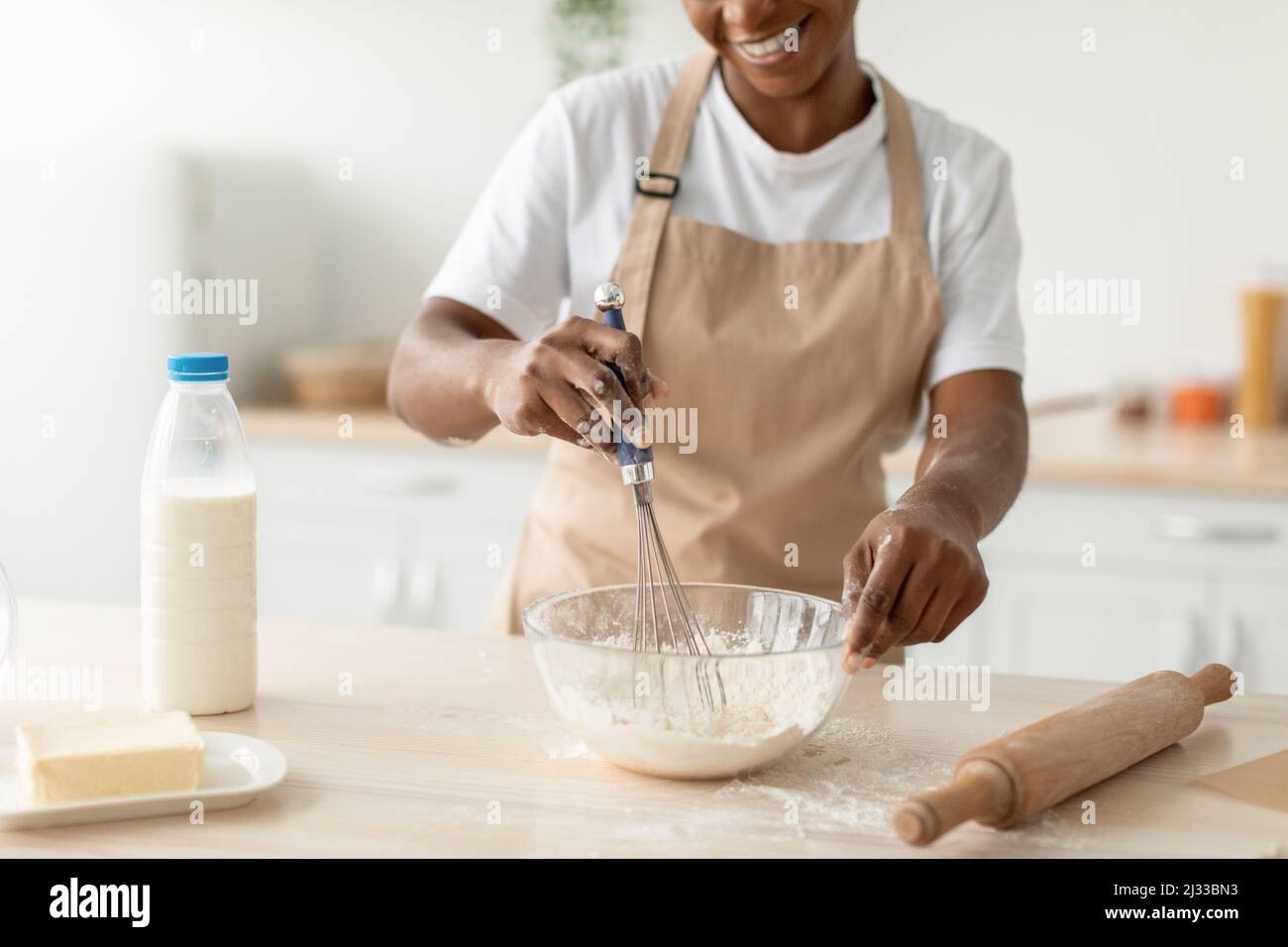 Smiling millennial african american woman in apron preparing dough for pie in minimalist kitchen interior Stock Photo