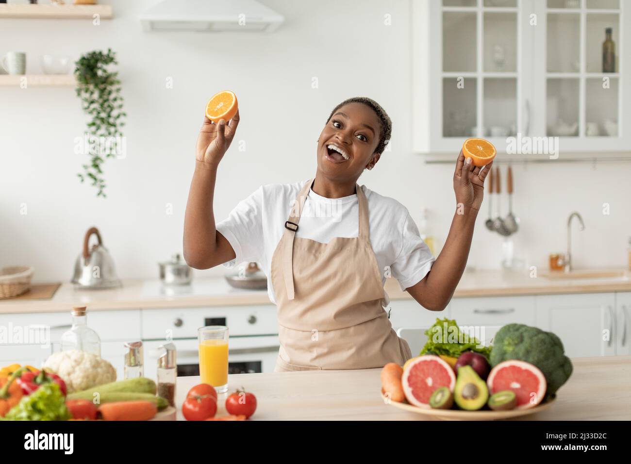 Glad emotional young black woman in apron have fun on minimalist kitchen interior, holds oranges Stock Photo