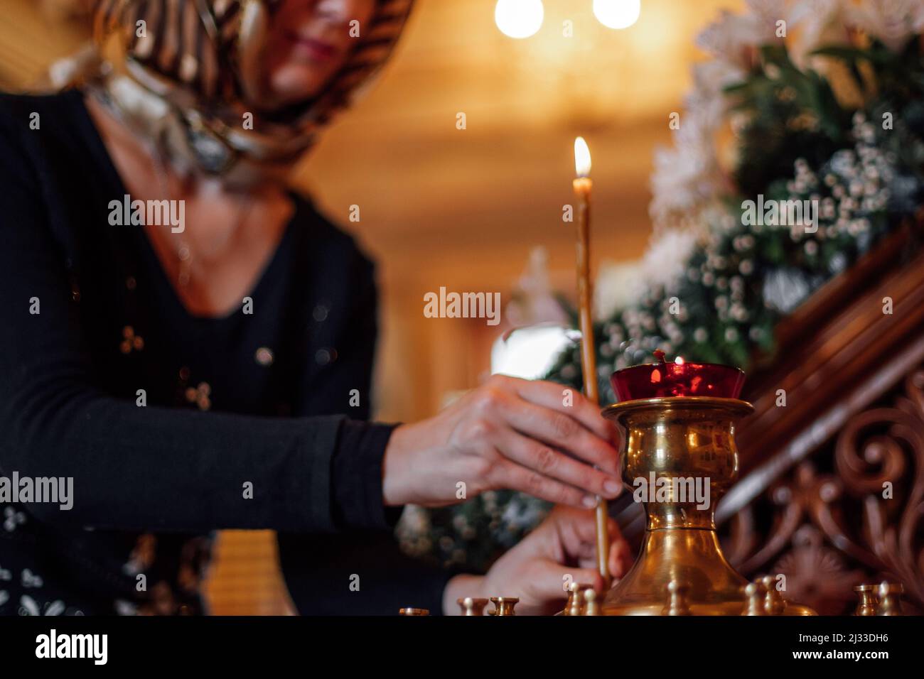 Cropped photo of woman wearing headscarf, putting burning candle on golden stand with candlesticks in orthodox church. Stock Photo