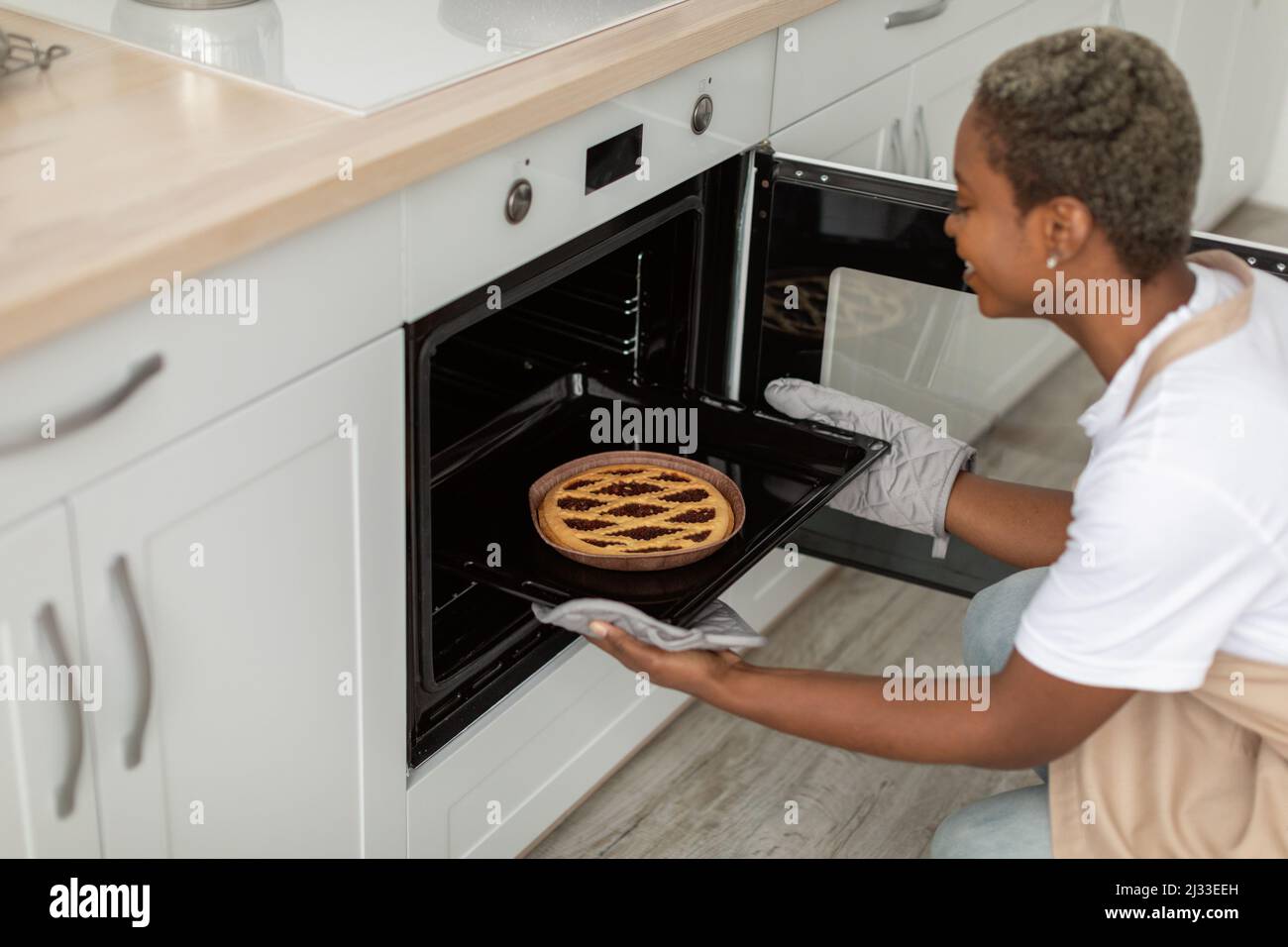Happy millennial african american female in apron takes out pie from oven in minimalist kitchen interior, profile Stock Photo