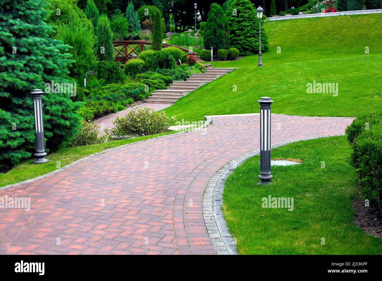 iron ground lantern and poli garden lighting of park curved path paved with stone tiles with drain lattice in park among plants, evergreen bushes and Stock Photo