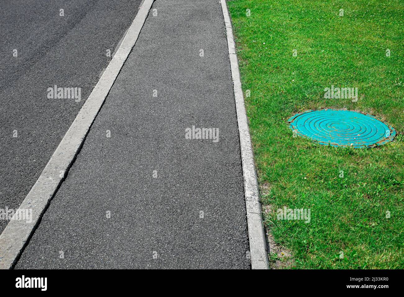 Asphalt road with tarmac footpath with stone curb and septic tank manhole on green lawn, nobody. Stock Photo