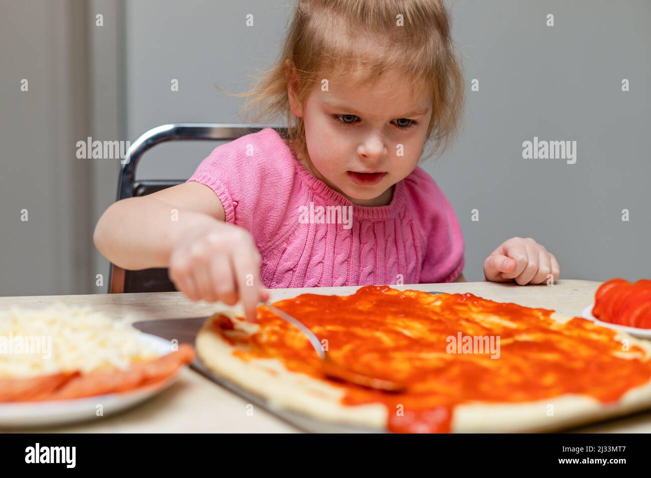 Small child cooking at home sitting at the table. Little girl making pizza in the kitchen. Kid learning to cook Stock Photo