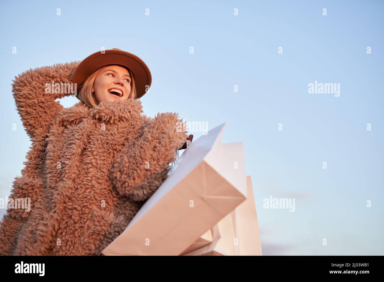 Excited happy caucasian fashion styled girl in hat and artificial fur outerwear standing amazed white shopping bags on blue sky background. Sale, consumerism, online shopping or black friday concept Stock Photo