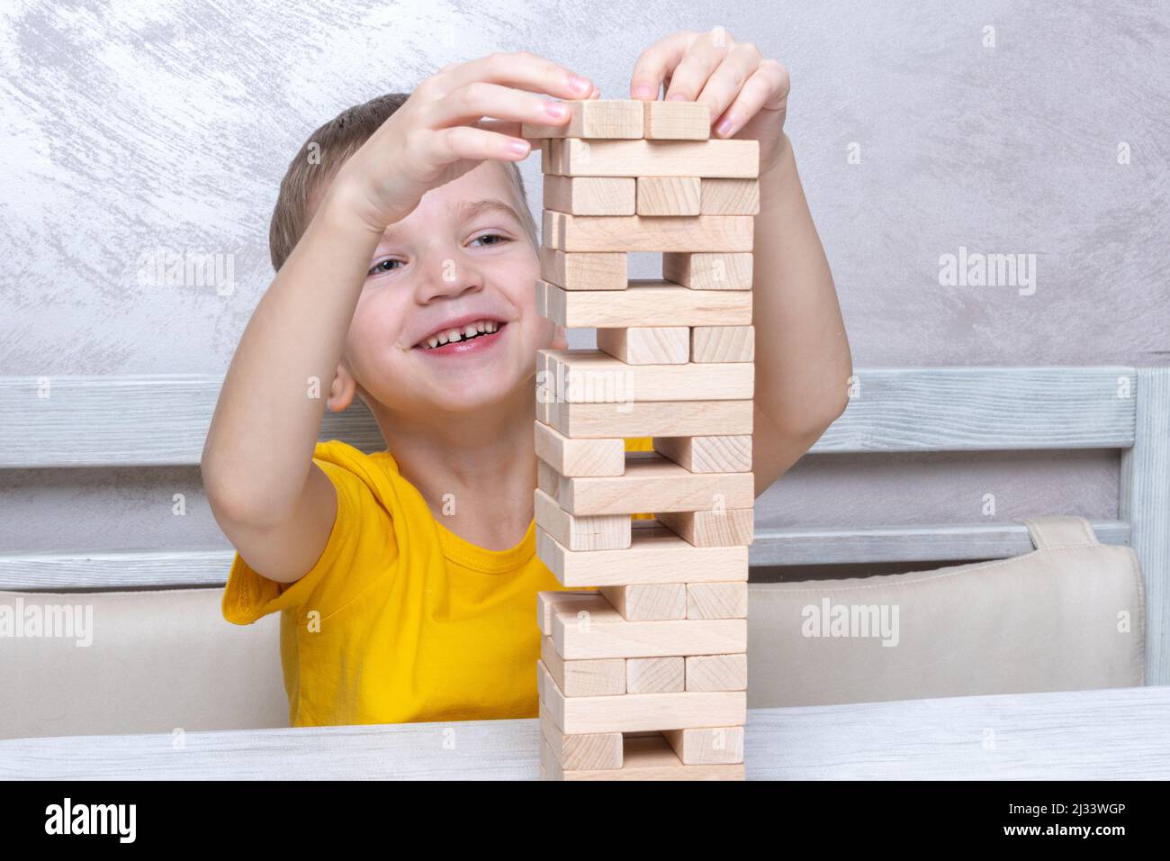 Interested happy little blond boy playing board game taking bricks from wooden tower keeping balance having fun together at home. Stock Photo
