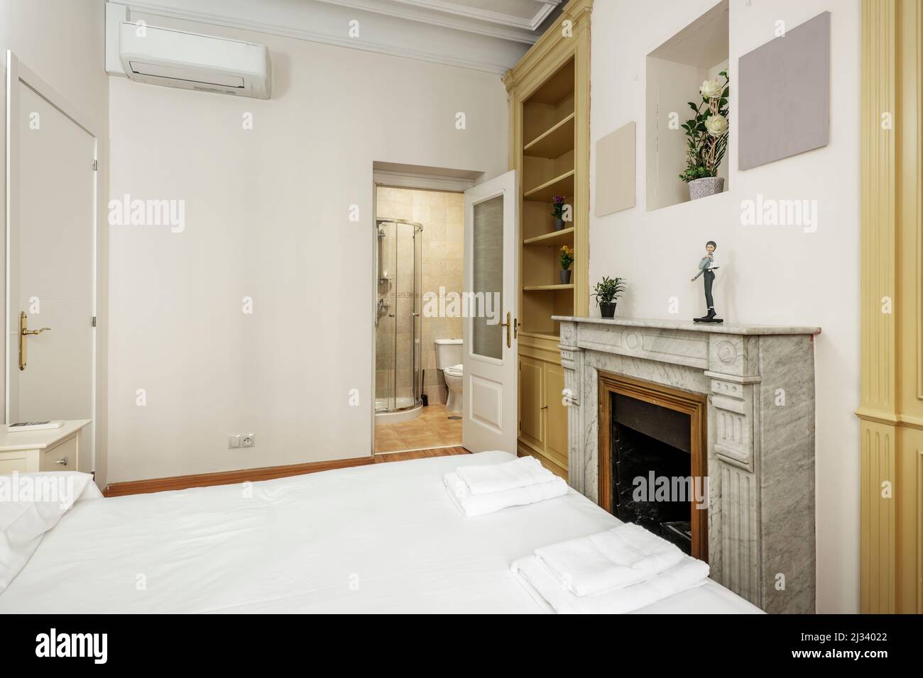 Bedroom with double bed, air conditioning, white marble fireplace, wooden shelves and shower cabin in an en-suite bathroom Stock Photo