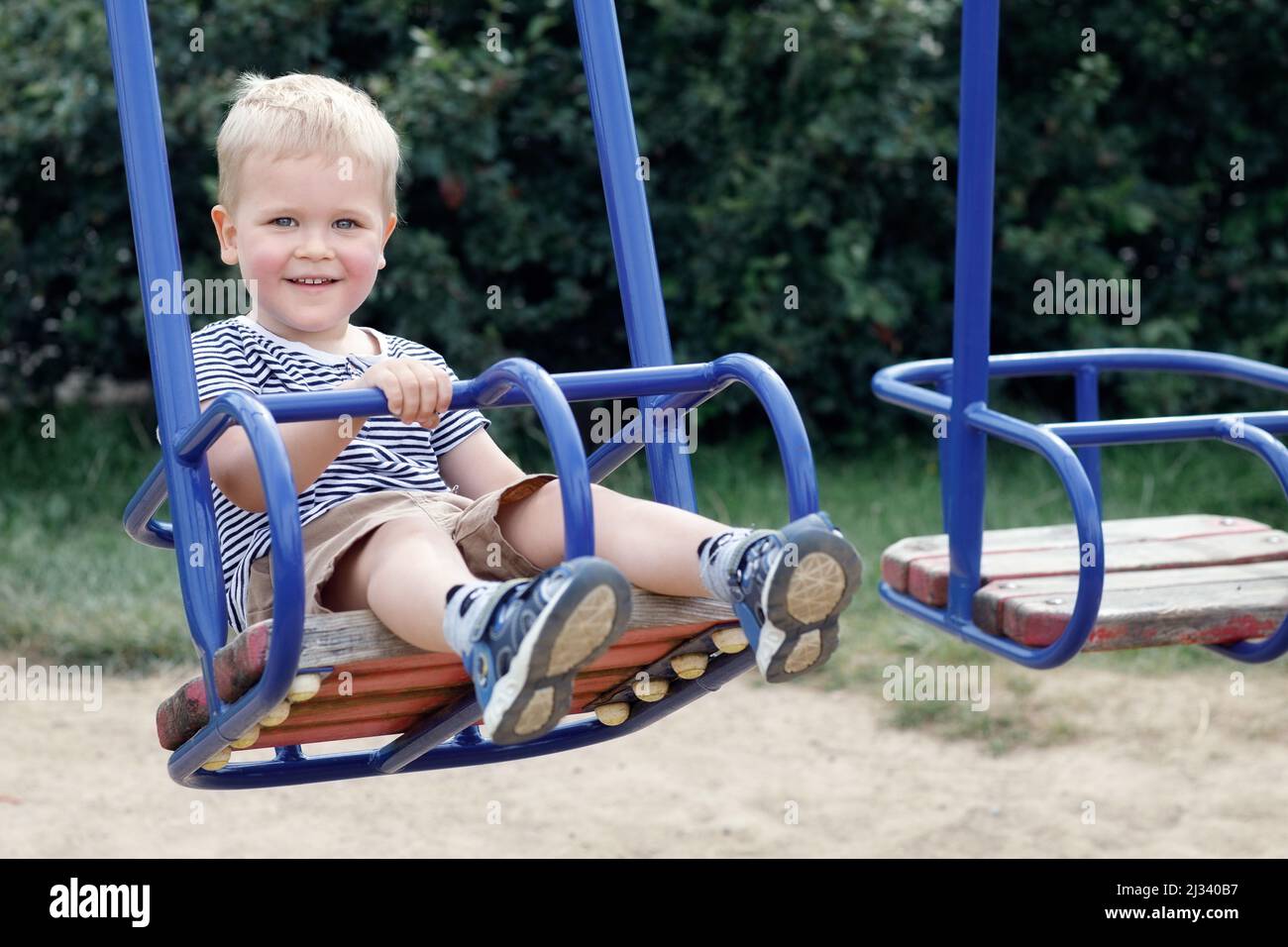 Little boy swinging on the swings of a public playground. Looking at camera and smiling. Happy childhood Stock Photo