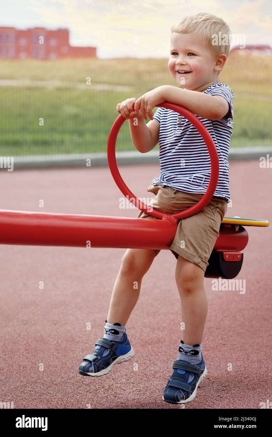 Beautiful boy sitting on a double sided swings at a playground. Smiling boy in summer time. Like to be raised. Happy kid in outside Stock Photo