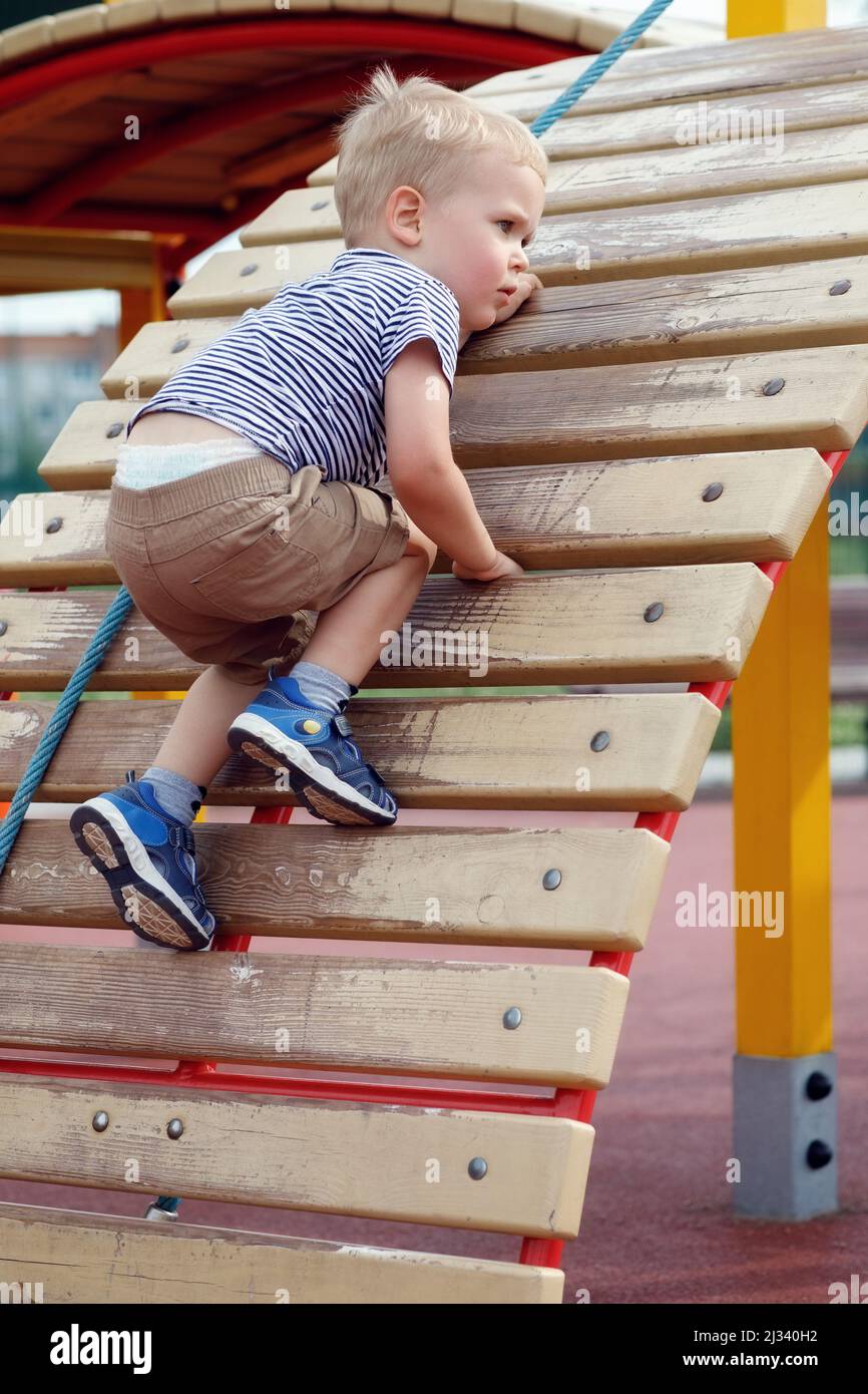 Anxious child trying not to fall down from climbing wall in a  playground. Strong and brave boy using an outdoor playground equipment Stock Photo