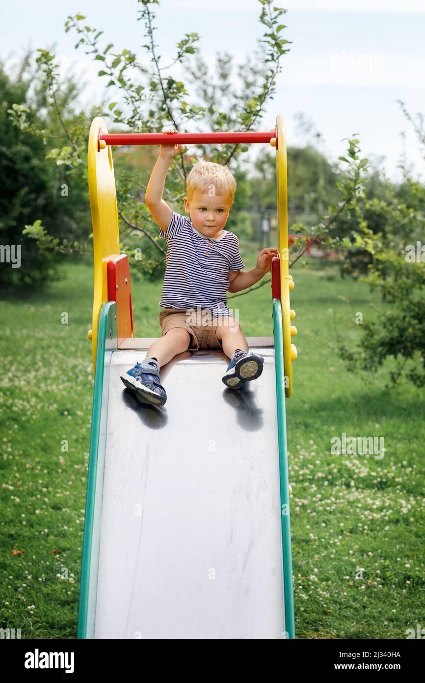 Little boy siting on a slide and contemplating to slide down. Green nature background. Childhood joys in summer time Stock Photo