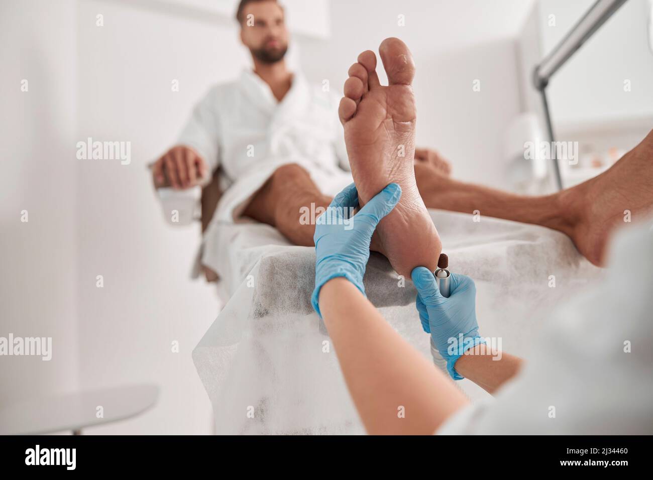 Beautician in gloves peels heel of male client with grinding tool in modern salon Stock Photo