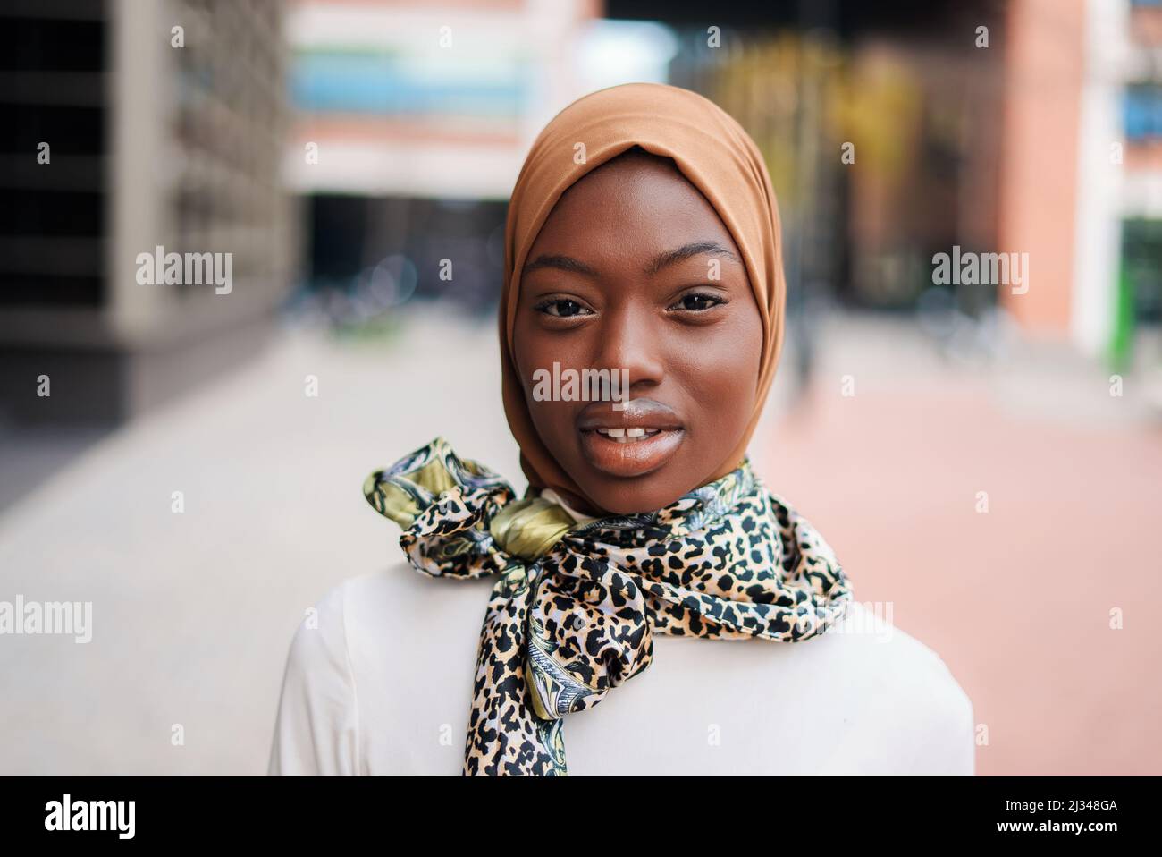 Charming Muslim African American female in traditional hijab and stylish scarf tied on neck standing on city street and staring at camera Stock Photo