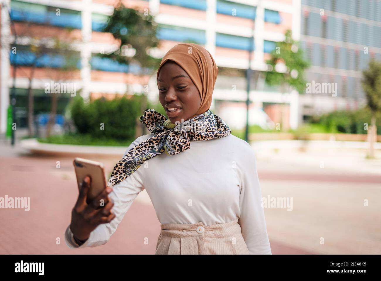 Positive young African American female in hijab reading message on cellphone on street in modern district Stock Photo