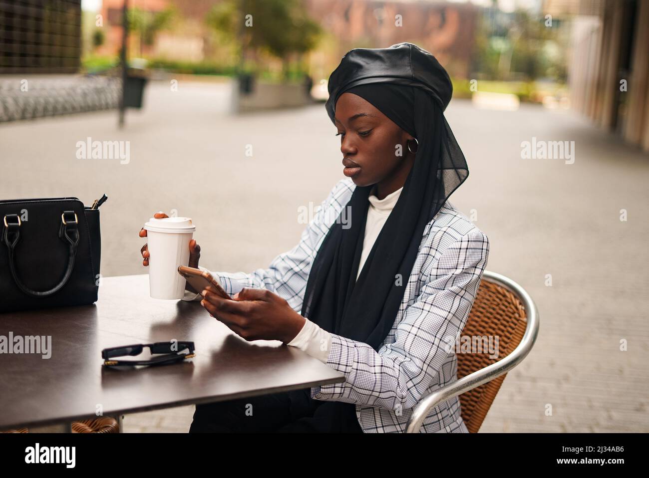 Serious African American female in trendy formal wear and traditional headscarf sitting at table and drinking takeaway coffee while surfing Internet on cellphone Stock Photo