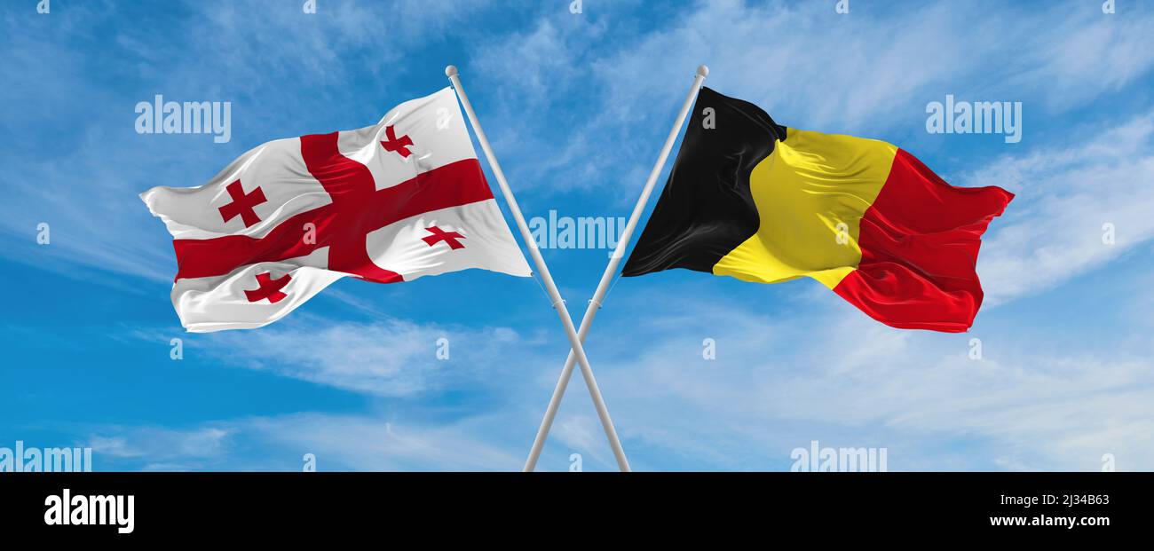 two crossed flags Georgia and Belgium waving in wind at cloudy sky ...
