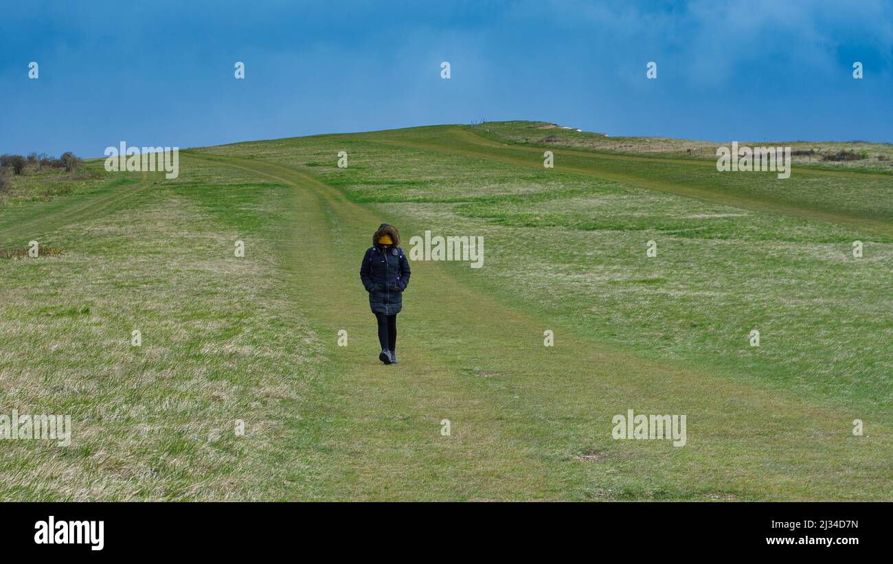 A beautiful hiker walking in the windy weather wearing a parka down jacket and hiking boots along beachy head cliffs, Eastbourne, United Kingdom Stock Photo