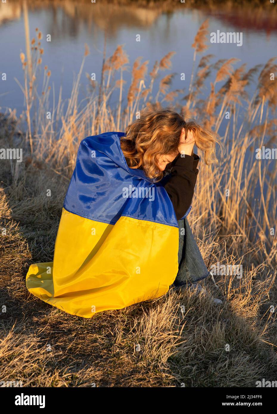 crying unhappy young patriot woman. wrapped in the Ukrainian flag sits on the shore of the lake in despair. war in Ukraine, Russian aggression. suffer Stock Photo