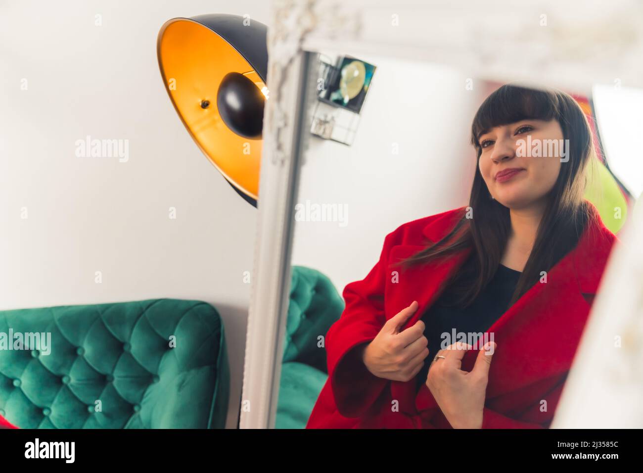 Young Caucasian dark haired woman trying on red coat. looking at the mirror portrait copy space . High quality photo Stock Photo