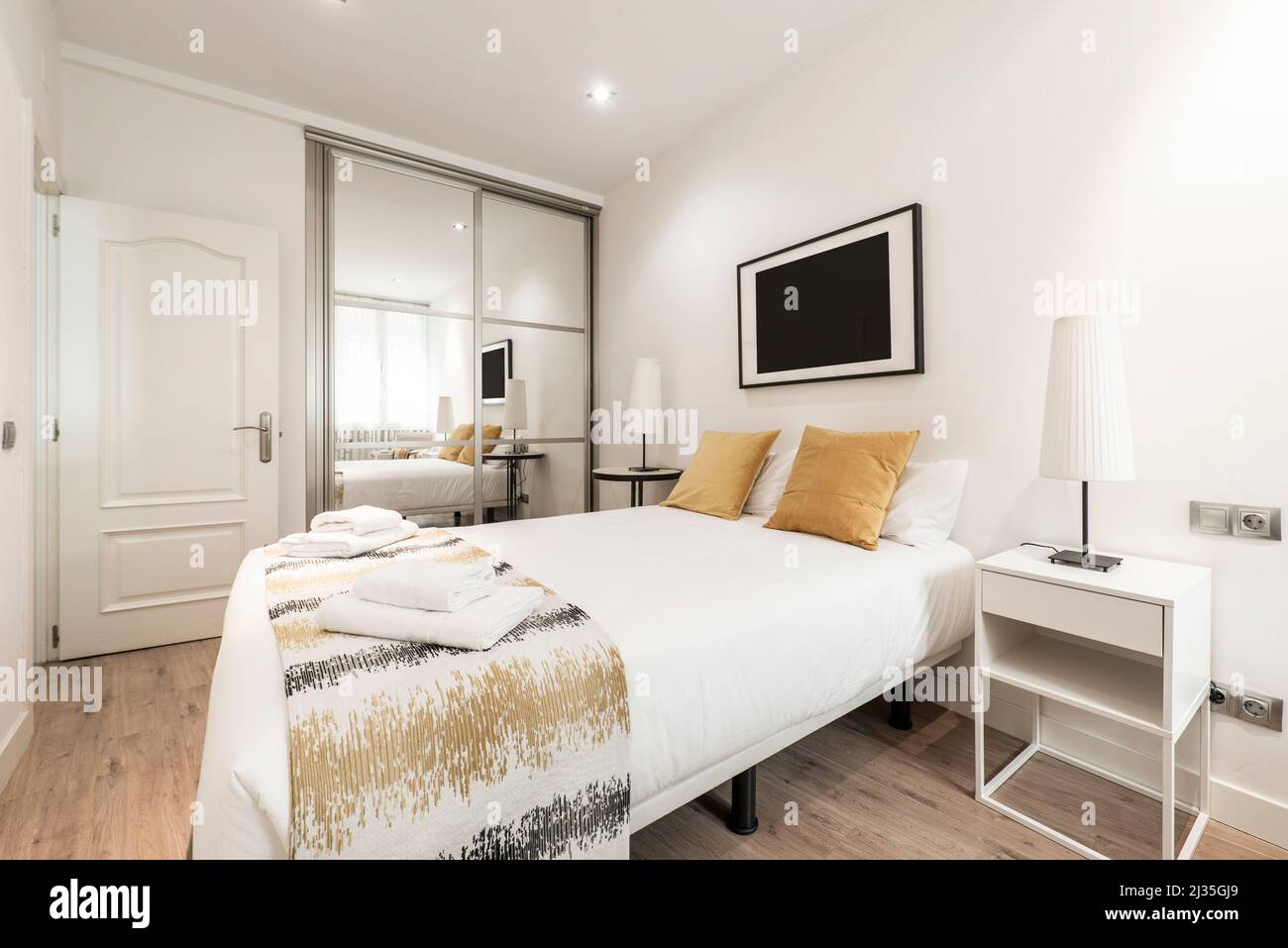 Bedroom with a large bed, gold cushions, a built-in wardrobe with sliding glass doors, uneven bedside tables and wooden floors in a short-term rental Stock Photo