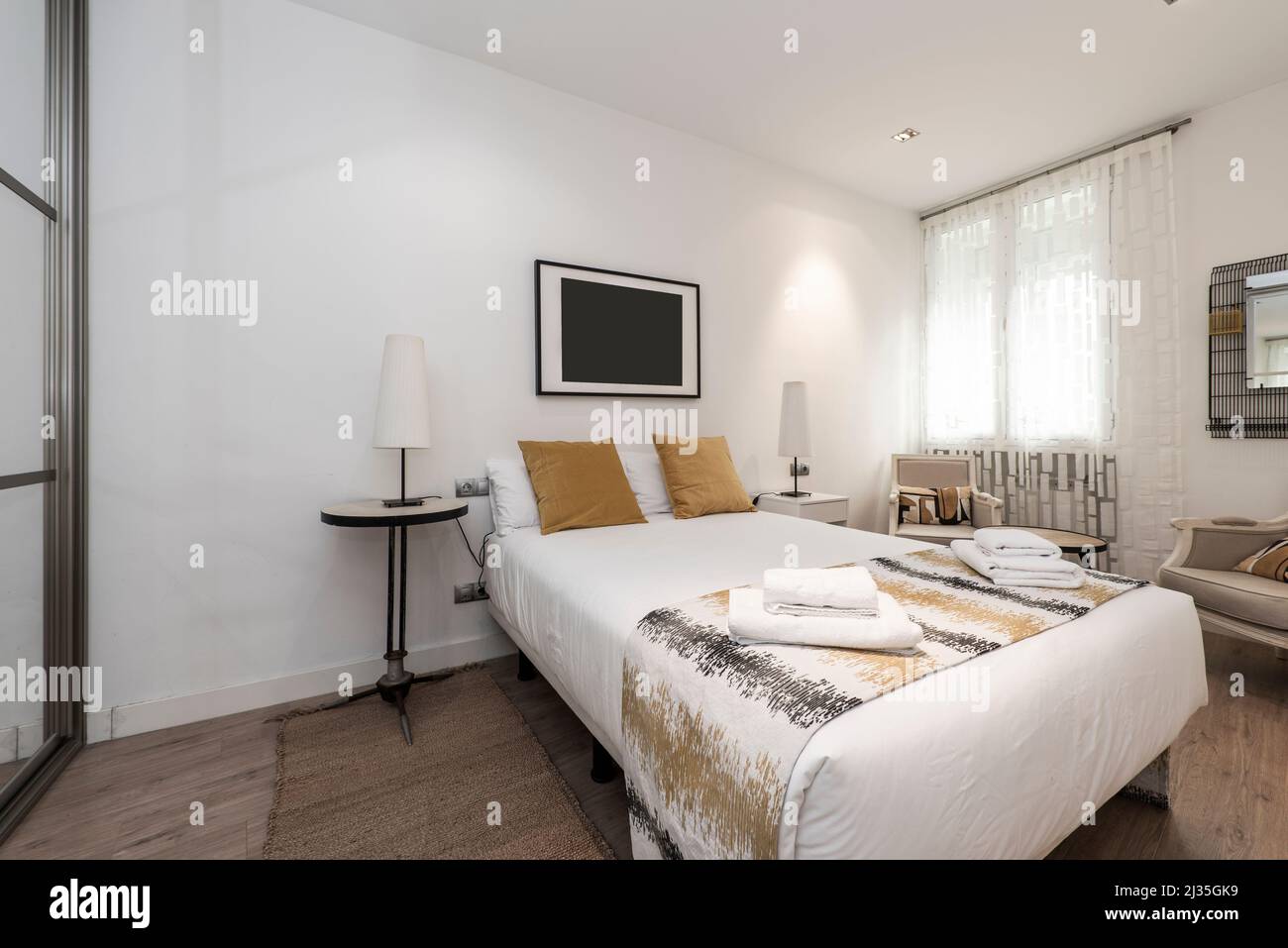 Bedroom with a large double bed with gold cushions, mismatched side tables, towels on the bed, and a wardrobe with mirrored sliding doors Stock Photo