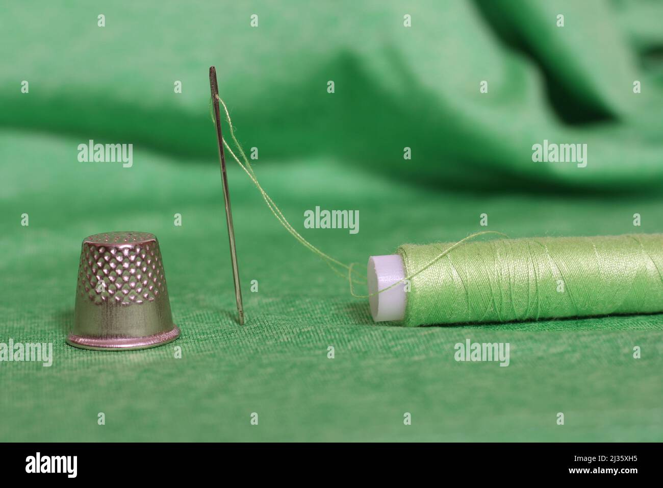 Spool of Green Thread, Thimble and Needle on Green Fabric Close up Stock Photo