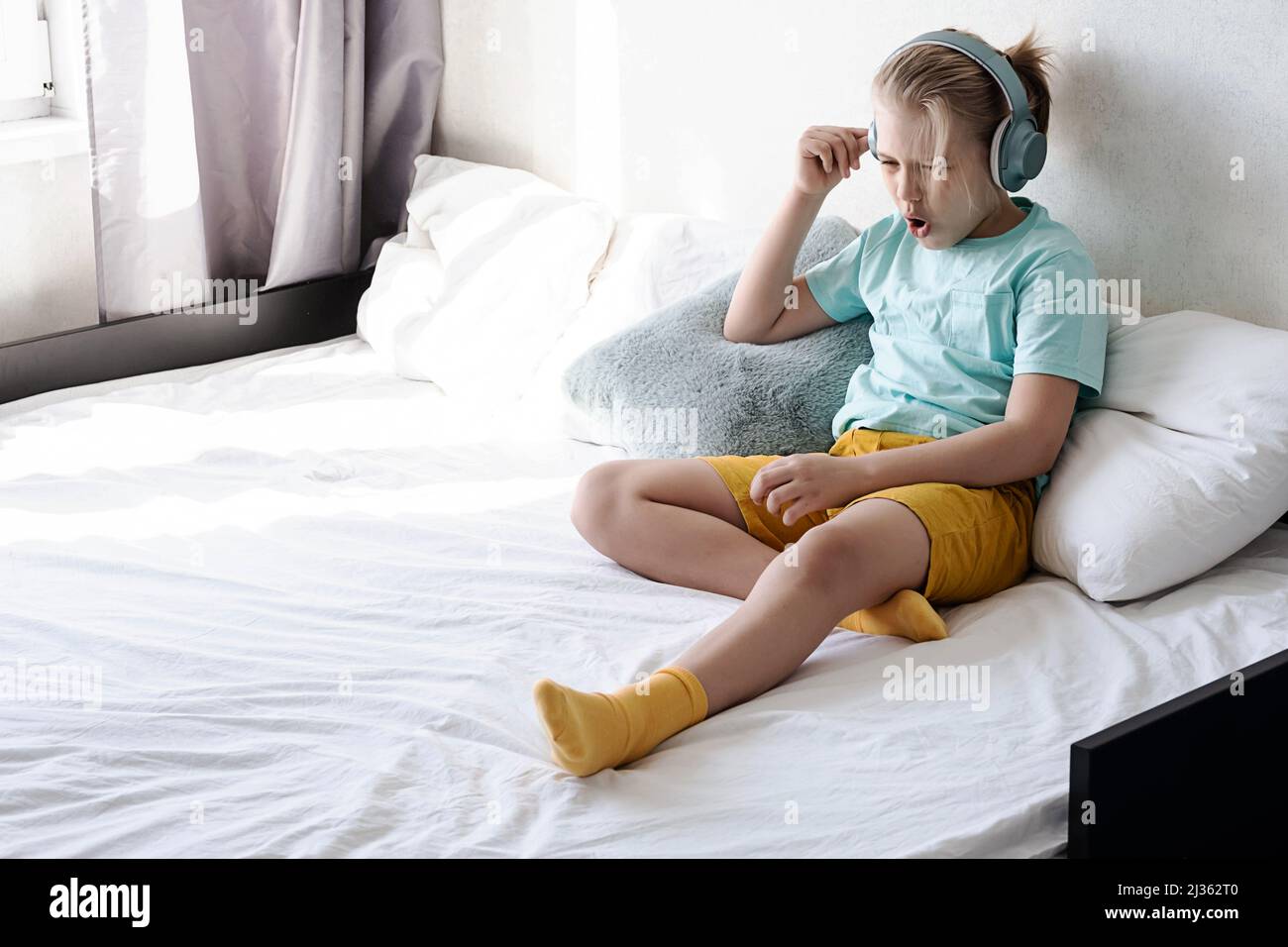 Child wearing headphones listens to music. Boy listening to music in headphones seating on bed. Stock Photo