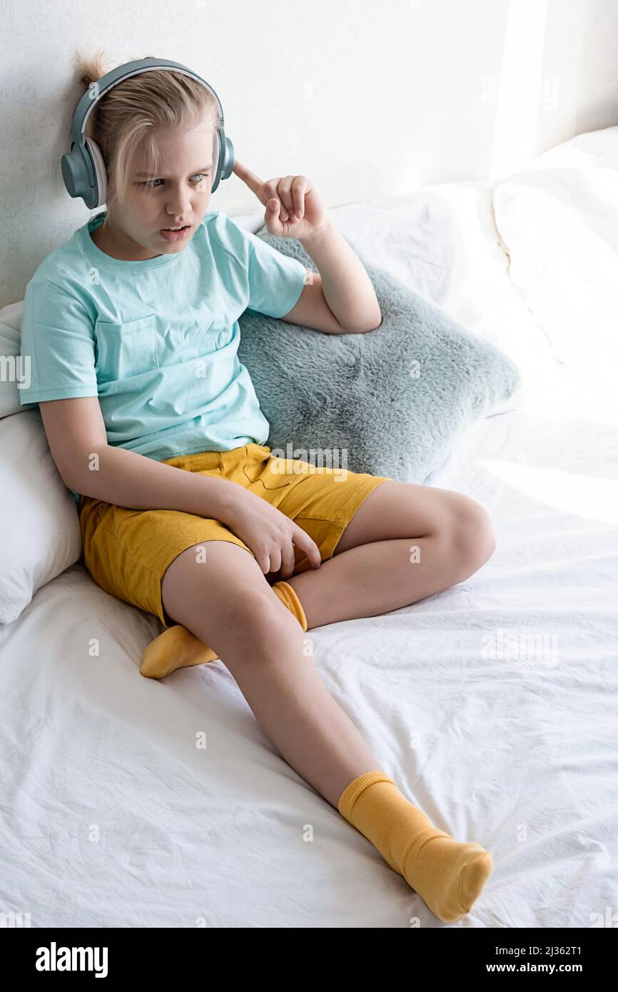 Child wearing headphones listens to music. Boy listening to music in headphones seating on bed. Stock Photo