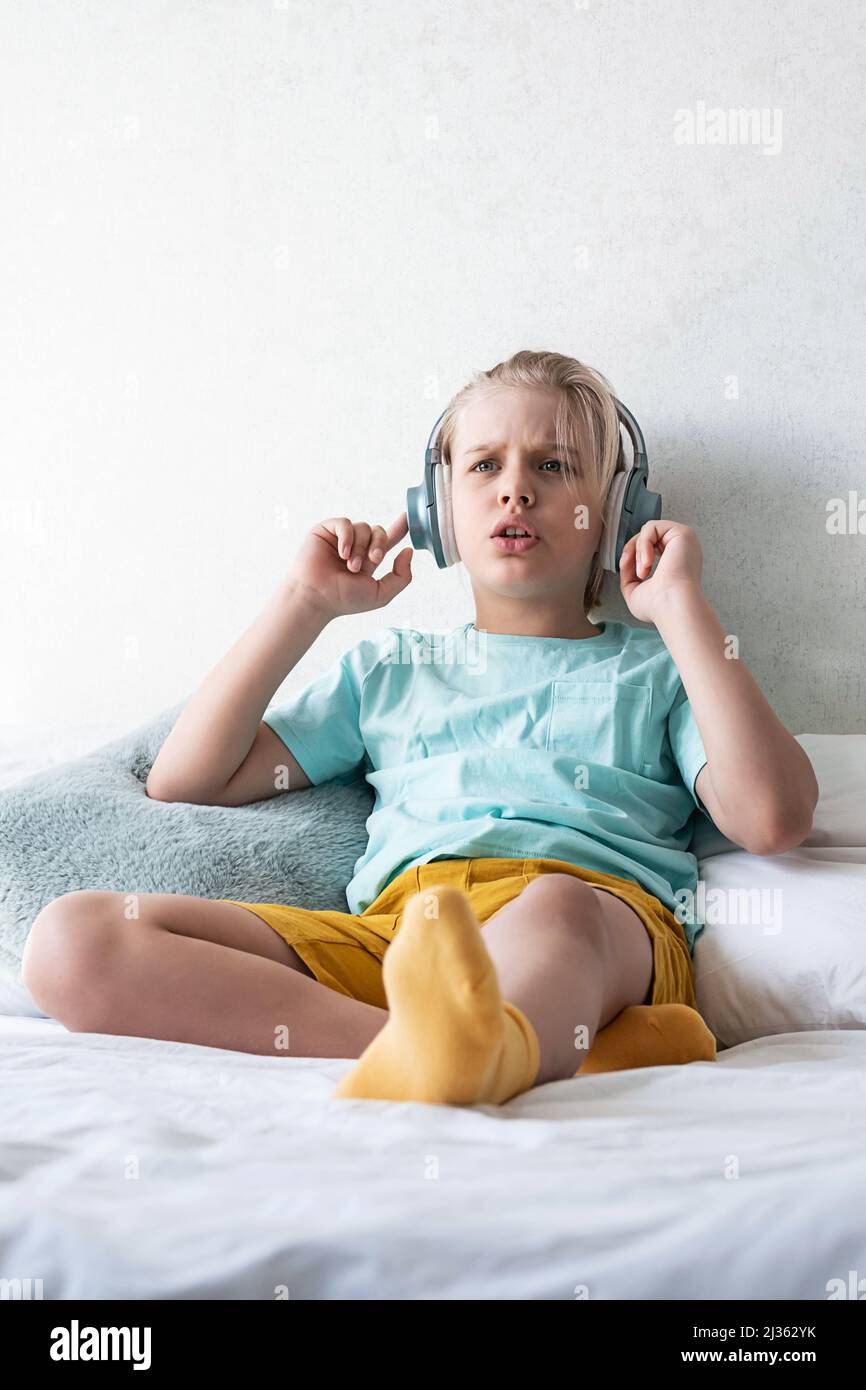 Child wearing headphones listens to music. Boy listening to music in headphones seating on bed. Stock Photo