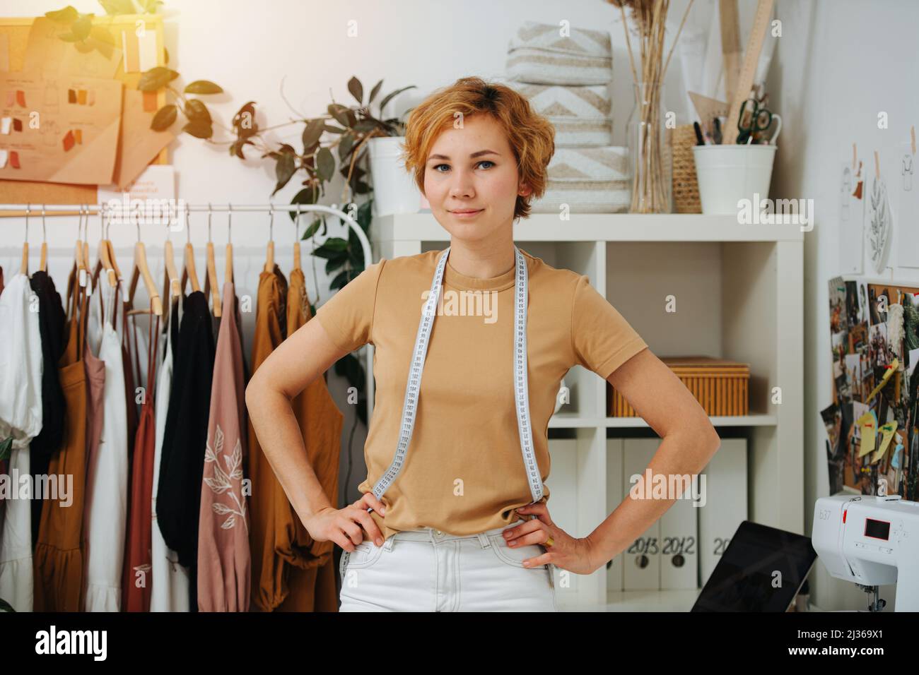 Proud seamstress posing for a photo in front of shelves and racks. Hands on hips. Meter hanging on her neck. She has short ginger dyed hair. Stock Photo