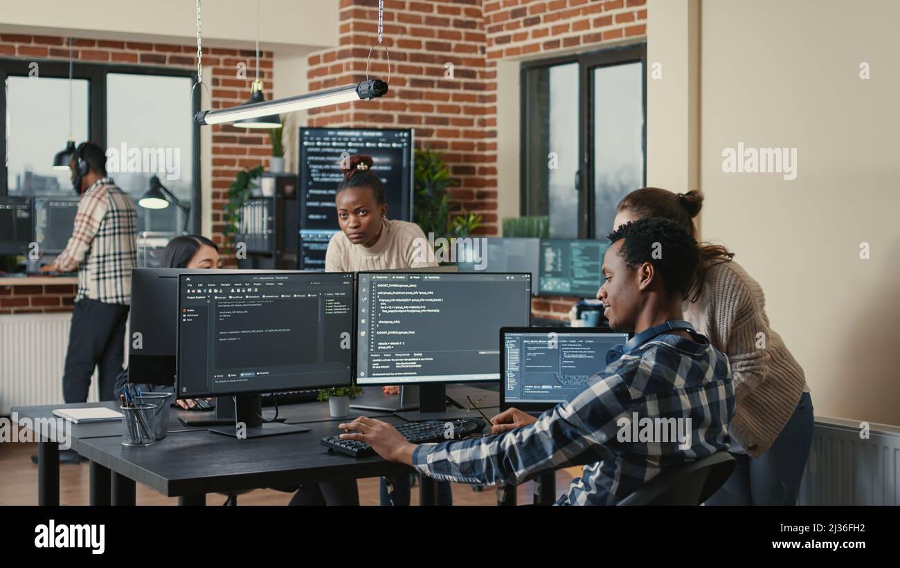 Programmer analyzing compiling code on multiple screens takes off glasses and doing high five hand gesture with colleague developer. Software developers celebrating successful coding. Stock Photo