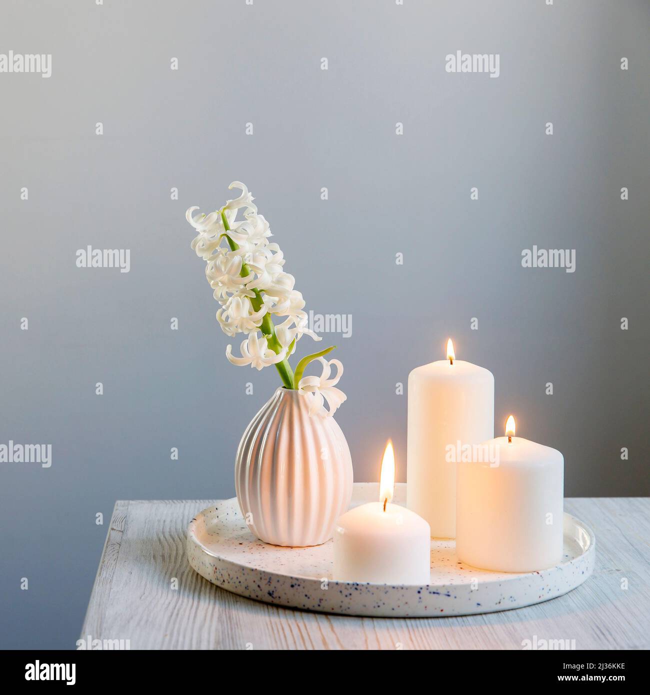 A bouquet of white cut hyacinth a in a small white corrugated vase and three large burning candles on a round tray are on a beige table. Place for tex Stock Photo