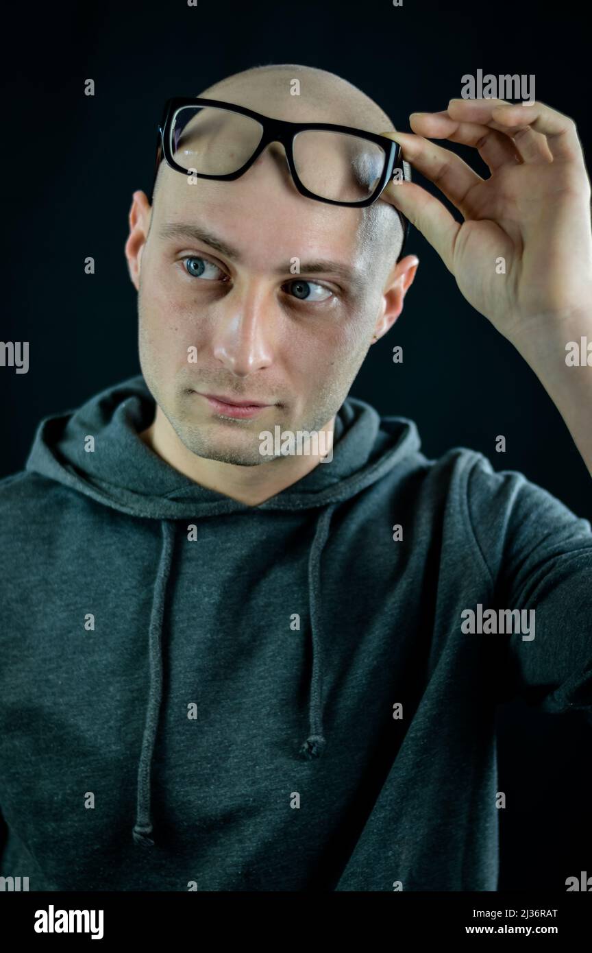 Portrait of a bald young man holding black eyeglasses on his forehead while looking at something to the side Stock Photo