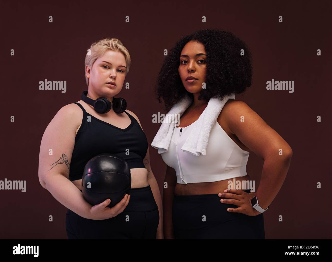 Two plus size women in sports clothes standing in studio. Young female athletes posing together against brown background. Stock Photo