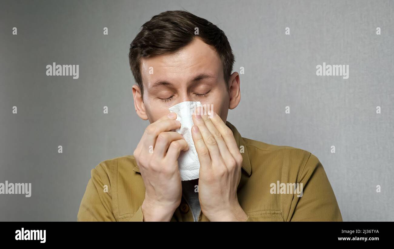 Sick man sneezes hard wiping with white napkin standing on grey ...