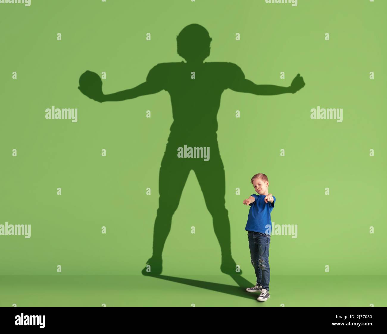 Conceptual image with little kid dreaming about future sport career and shadow of male american football player on green background. Stock Photo