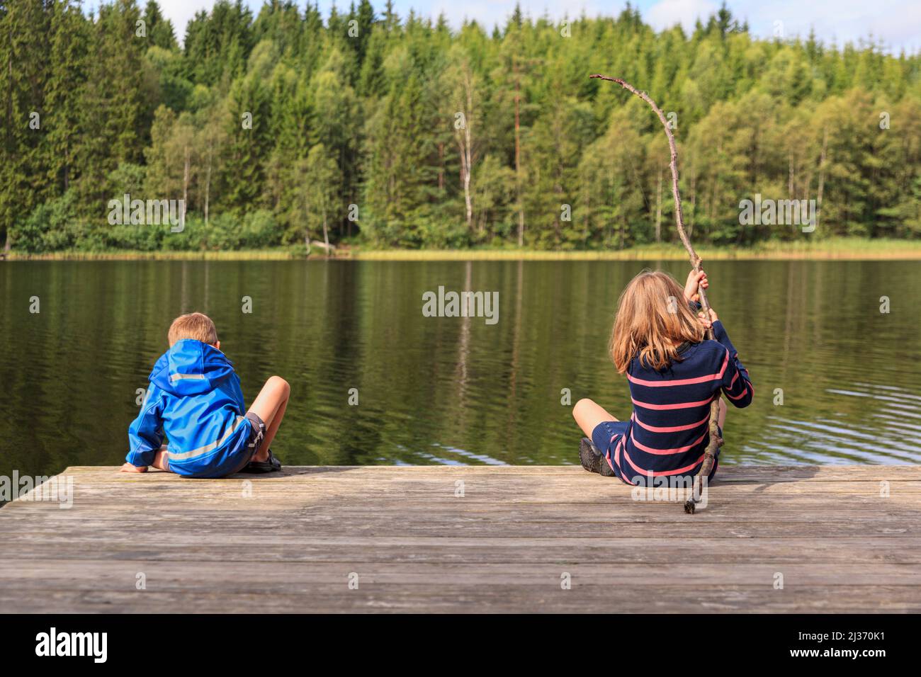Two children, younger brother and older sister, using a homemade fishing rod fishing from jetty by a lake set in an idyllic Swedish summer forest landscape Stock Photo